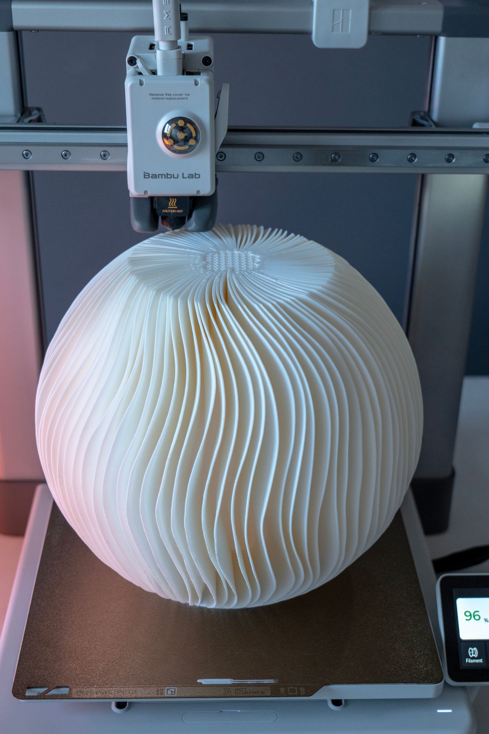 A large white lampshade of wavy vertical fins nearly finished on the A1's build plate, under the print head.
