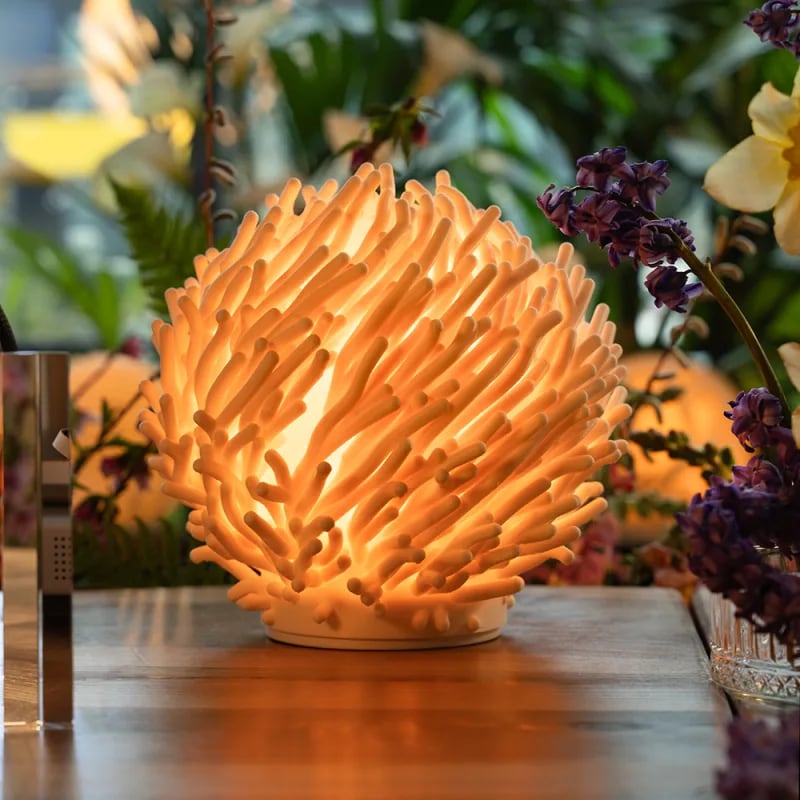 A Coral lamp glowing warm on a wooden table beside purple flowers and green leaves.