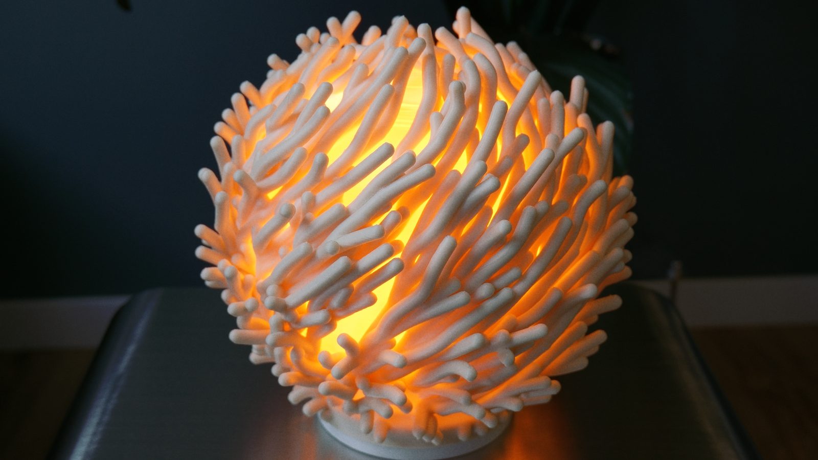 A 3D printed lampshade of branching, coral-like tubes in natural PHA, glowing warm orange from the light inside.